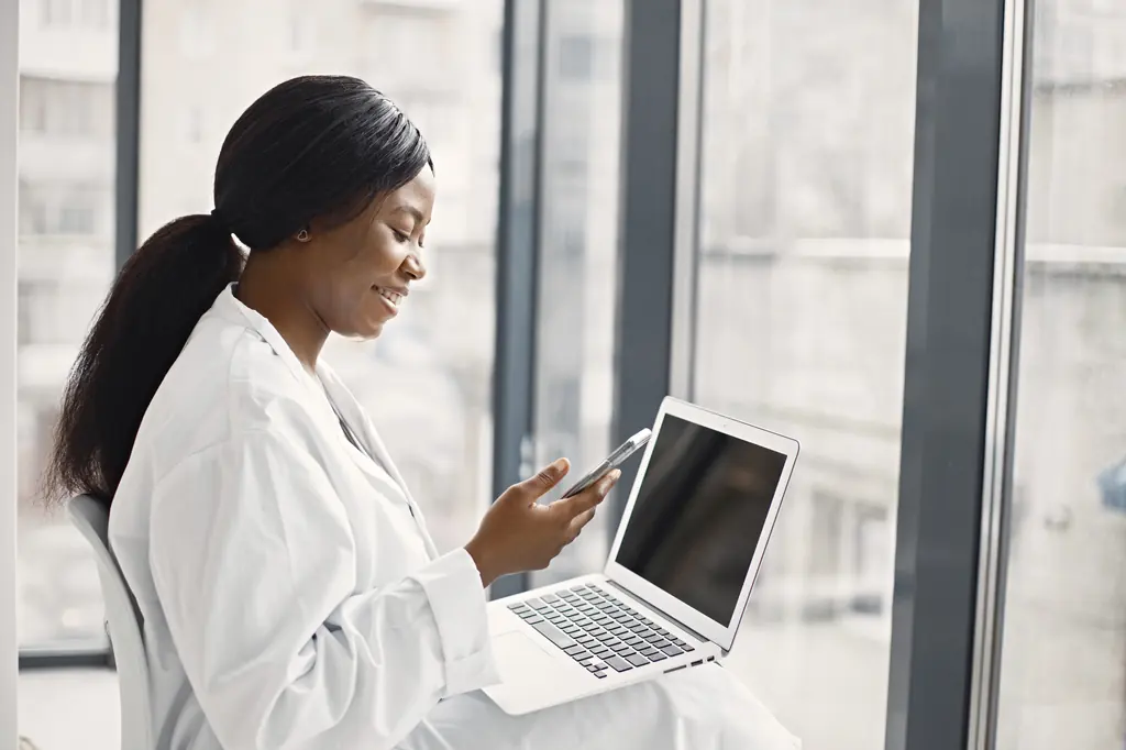 A doctor by a window, checking her phone with a laptop on her knees